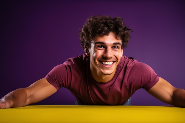 A man in a red shirt smiles with his arms outstretched and his face is leaning on a yellow table.