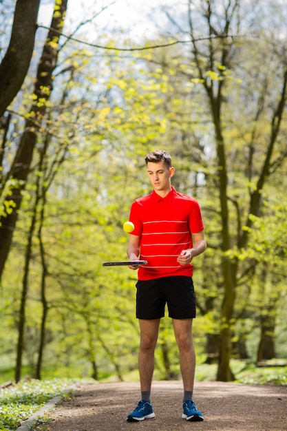 赤いtシャツの男は、緑豊かな公園の背景にテニスラケットとボールでポーズします。