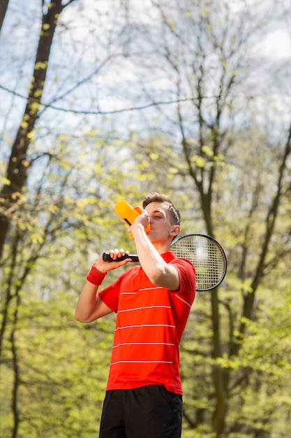 The man in a red t-shirt pose with a tennis racket and drinking water from a thermocouple. Sport concept