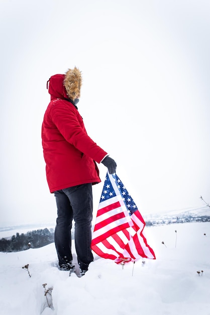 Мужчина в красном зимнем пальто держит флаг сша на улице в снежную погоду