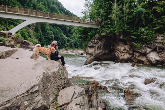 Мужчина отдыхает у горной реки, сидя на камне. Рядом бежит коричневая собака.