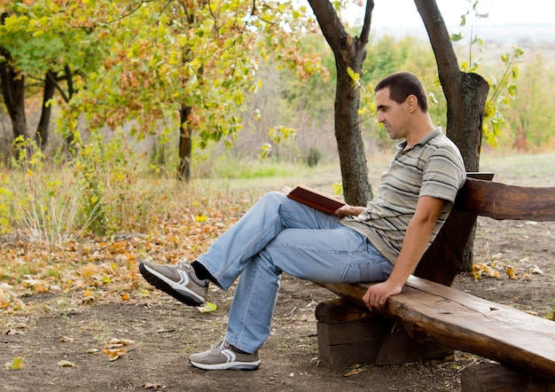 Man relaxing with a book sitting on a rustic wooden bench with a woodland backdrop as he relaxes and enjoys his vacation