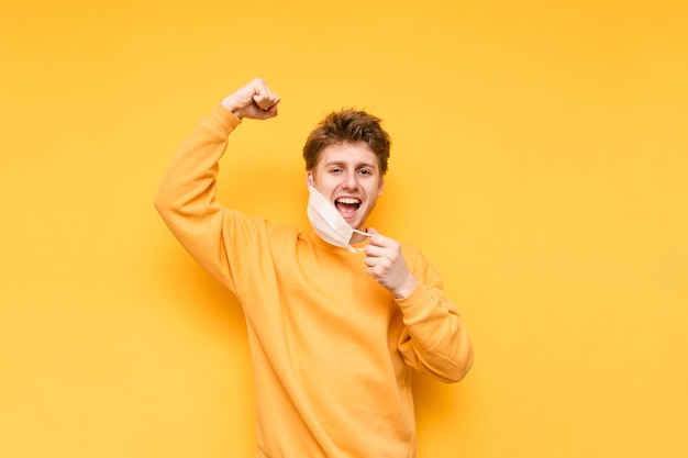 man removes a medical mask from his face on a yellow background looks at the camera and rejoices