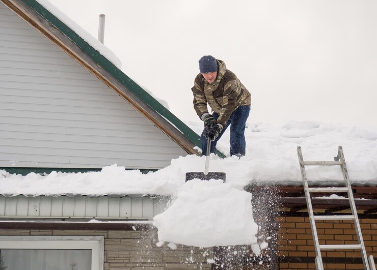 A man removes snow from the roof of a completely snowcovered house with a shovel a lot of fresh snow after a blizzard hard and dangerous work