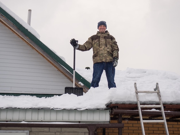 Мужчина убирает снег с крыши полностью заснеженного дома лопатой много свежего снега после тяжелой и опасной работы вьюги
