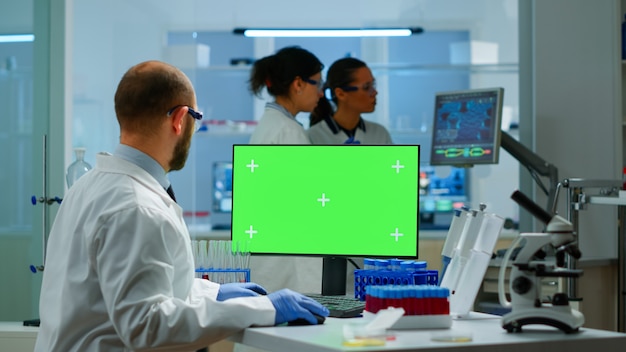 Ricercatore dell'uomo che guarda il display della chiave di crominanza nel moderno laboratorio attrezzato digitando sul computer. team di microbiologi che effettuano ricerche sui vaccini scrivendo su dispositivo con schermo verde, isolato, display mockup.