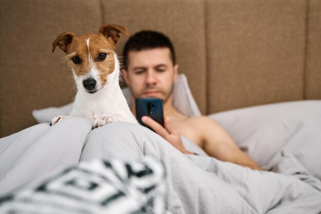 犬と一緒にベッドで休んでスマートフォンを使用する男性