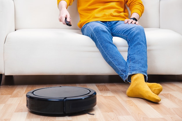 Man resting on the sofa, while the robot vacuum cleaner cleaning living room and dust. Concept time for yourself. Man controlling vacuum cleaner with remote control