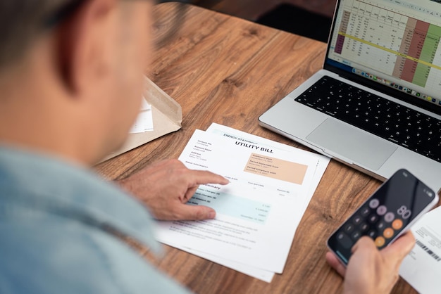Man reviewing utility bills at home while using a smartphone calculator Personal finance management and budgeting concept Ideal for household expenses and tax calculation themes