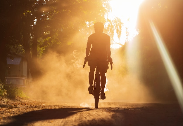 夕暮れ時に砂利道で自転車に乗る男。ほこりの雲の中の砂利自転車のサイクリストのシルエット。