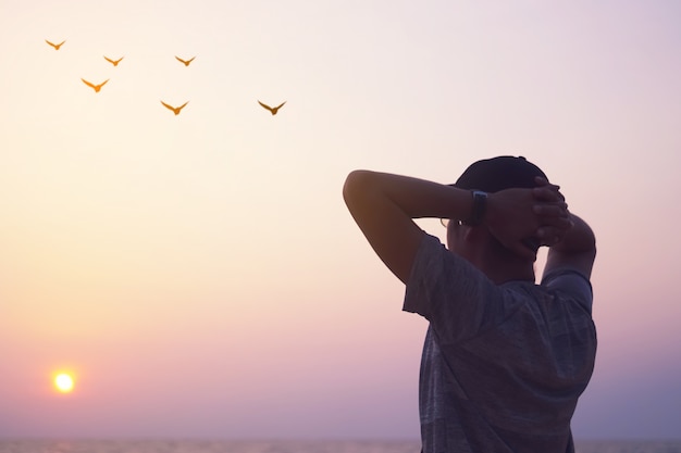 L'uomo aumenta le mani fino al cielo che esamina gli uccelli volare attraverso il concetto di libertà della metafora con il cielo del tramonto e il fondo della spiaggia dell'estate.