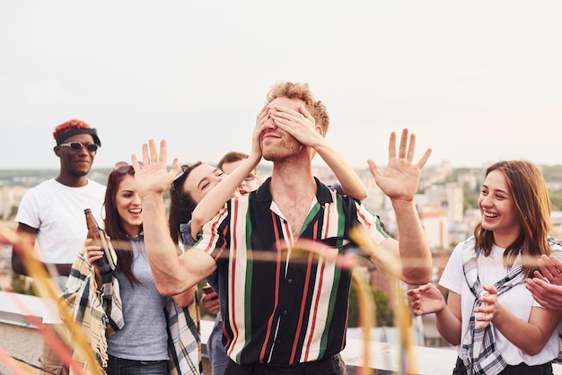 Man's eyes covered by hands Group of young people in casual clothes have a party at rooftop together at daytime