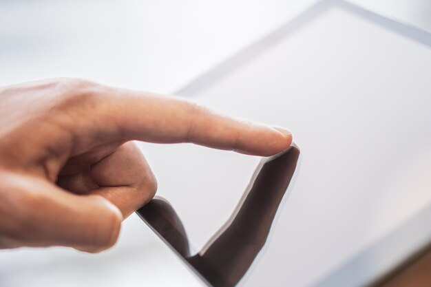 Photo man's finger touching a digital tablet screen placed on a desk surrounded by the vague surroundings of a bright office setting