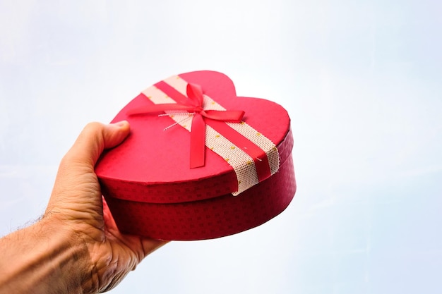Мужская рука дарит подарок в коробке в форме сердца с бантом, на белом фоне.