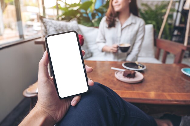 man's hand holding black mobile phone with blank white screen with woman sitting in cafe