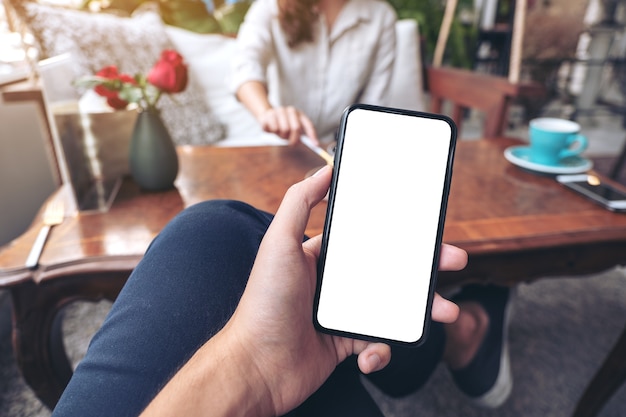 man's hand holding black mobile phone with blank white screen with woman sitting in cafe