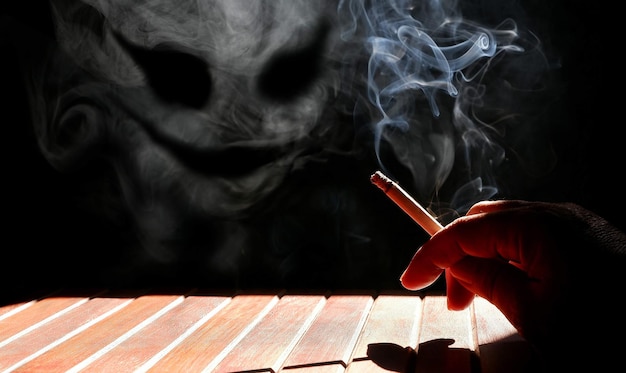 Man's hand holding cigarette with smoke look like devil face floating on the air in dark background