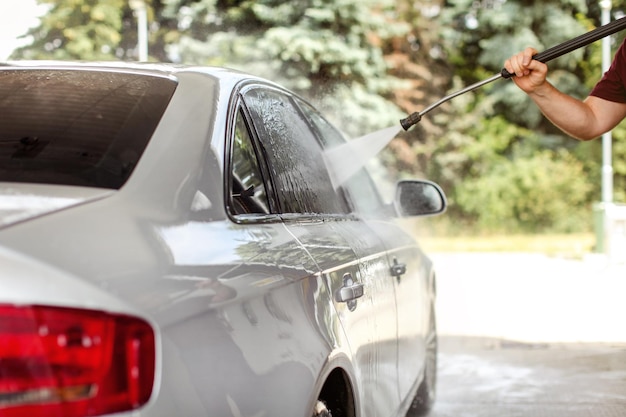 Man's hand holding hose, spraying water to widow of silver car in self serve carwash, sun lit blurred green trees in background.