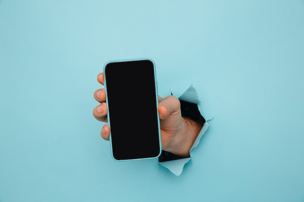 Man's hand holding phone from blue torn paper.