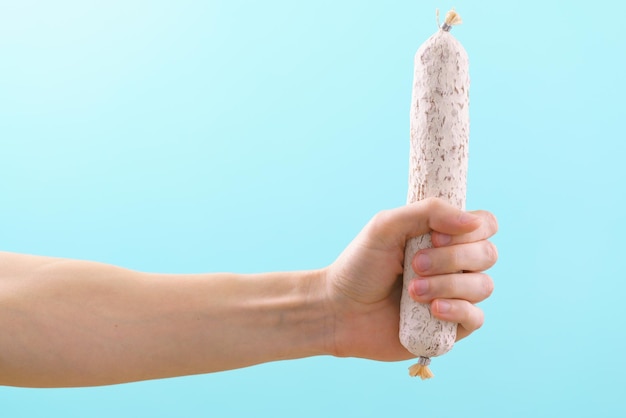 Man's hand holding a solid white Spanish fuet sausage on a blue background Sausage Spanish Hand Old Background Food Spain Mexican Thin Buffet Fat Tripe Pork Spicy Appetizer