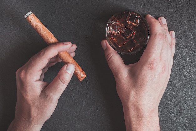 a man's hand holds a cigar and a glass of whiskey with ice