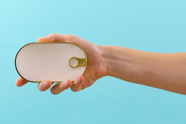 The man\'s hand holds a closed golden rounded empty fish tin can\
with an empty white paper mockup on a blue background sardine\
container can fish package ring nutrition closed canning close