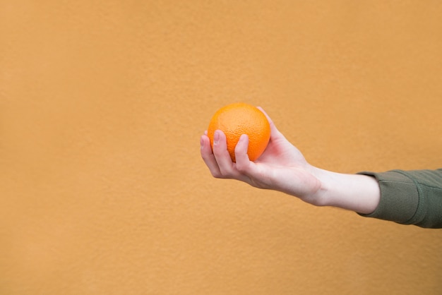 Man's hand holds an orange on an orange wall. Concept is a man and a fruit