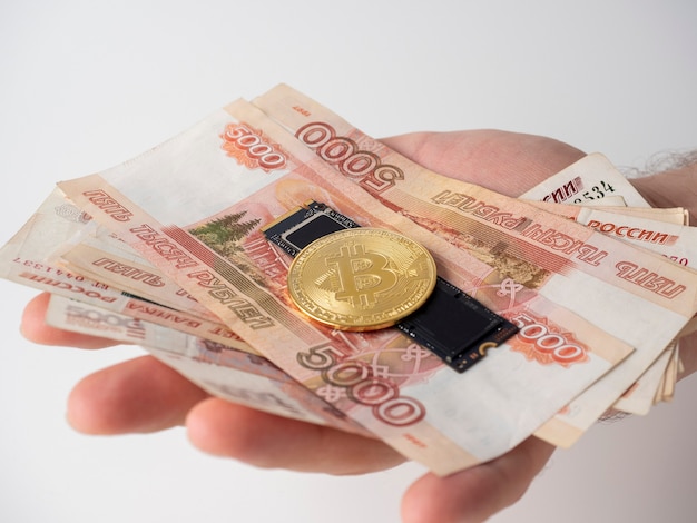 A man's hand holds Russian banknotes, an m2 SSD disk and bitcoin. The concept of earning and mining money on hard media