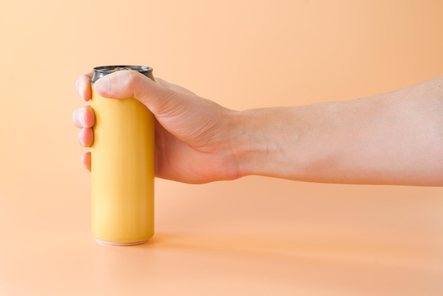 Мужская рука держит желтую алюминиевую банку пива или энергетического напитка, стоящую на оранжевом фоне.