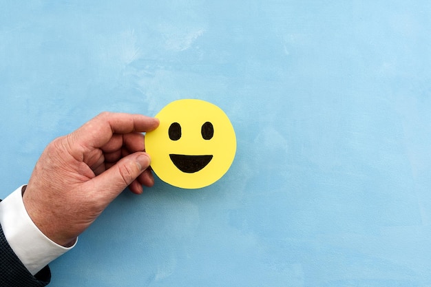 La mano dell'uomo tiene una faccia gialla con un'icona a faccia felice su sfondo blu il concetto di soddisfazione del cliente o feedback positivo affari e feedback