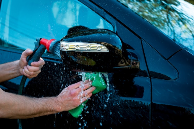 Man&#39;s hand polijsten van zwarte auto door spons en een andere hand hand houdt de slang voor het wassen buiten.