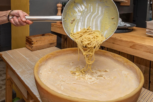 Man's hand pouring fat spaghetti from a frying pan over half grana padano cheese in an Italian restaurant