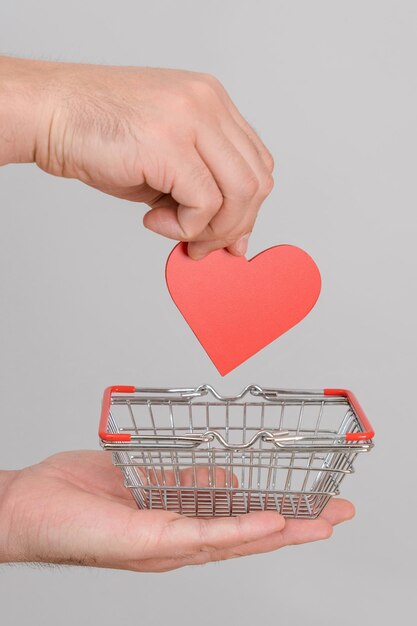 A man's hand puts a red heart symbol in a miniature shopping basket. The concept of love for money