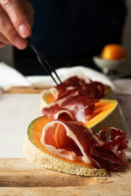 Foto mano d'uomo che serve, prova, assaggia, dispone le tradizionali tapas spagnole jamon iberico con melone e basilico su un tagliere di marmo.