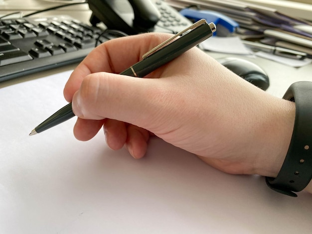 A man's hand in a shirt and with a fitness bracelet holds a pen and writes on the table