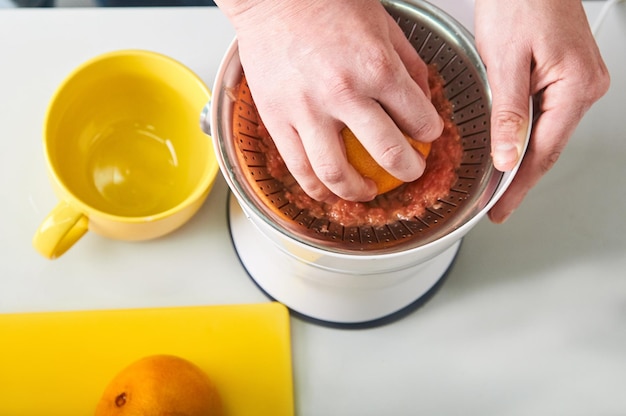 мужская рука выжимает грейпфрутовый сок на белой соковыжималке кухонная утварь и фрукты на столе вид сверху