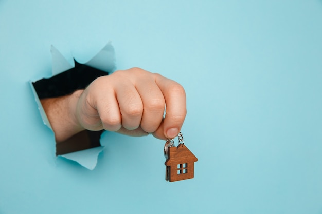 青い紙のクローズアップの穴を通して家の鍵を持つ男の手。住宅販売と家賃のコンセプト。