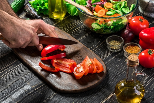 Man's hands cutting red paprika with knife. Cook cut red paprika. Man loves cooking fresh salad for dinner. Paprika cut by cook's hand. Close up of cutting board and vegetables