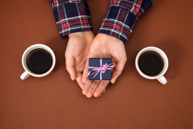 Man's hands keep box gift near two cups of coffee on brown.