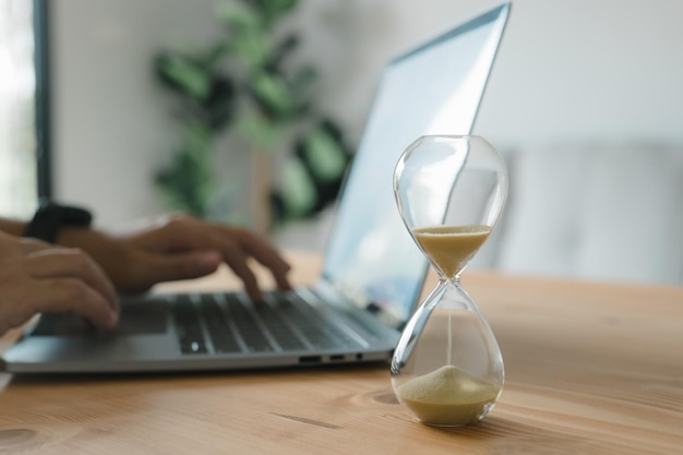 Man's hands typing on a laptop keyboard next to an hourglass Concept of time management business schedule and deadline