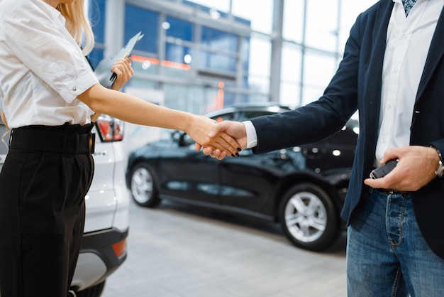 Man and saleswoman shake hands in car dealership. Customer and seller in vehicle showroom, male person buying transport, auto dealer business