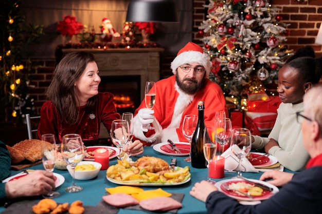 Uomo in costume da babbo natale che augura buon natale a tavola festiva, proponendo un brindisi, tenendo in mano un bicchiere di spumante. celebrazione delle vacanze invernali con la festa della famiglia a casa, raduno di amici diversi