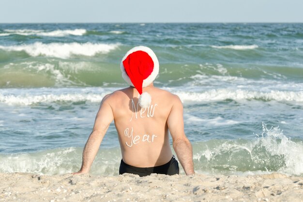 Man in Santa hats with the inscription New Year on the back on the sea beach. Back view