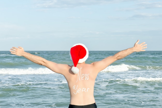 Человек в шляпах Санты с надписью Новый год на спине на берегу моря. Руки вверх. Вид сзади