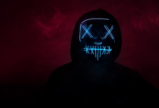 A man in a scary neon glowing mask and a hood on a dark wall with smoke. Horror and Halloween concept