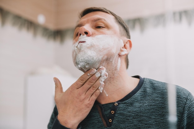 A man shaves his beard in front of a mirror in his bathroom.