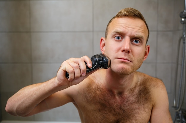 A man shaves his face with an electric razor in front of a mirror. Skin irritation. Bath procedure