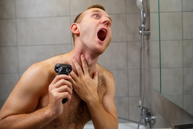 A man shaves his face with an electric razor in front of a mirror. Skin irritation. Bath procedure