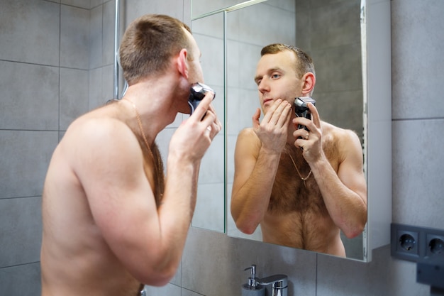 A man shaves his face with an electric razor in front of a mirror. Skin irritation. Bath procedure