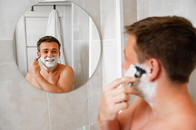 A man shaves his face with a safety razor and looks in a round mirror